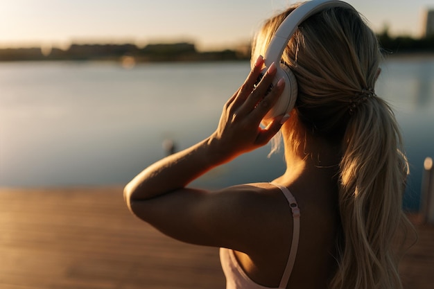 Widok z tyłu młodej wysportowanej blondynki w sportowej odzieży treningowej zakładającej bezprzewodowe słuchawki, aby zacząć biegać w parku miejskim w letni poranek