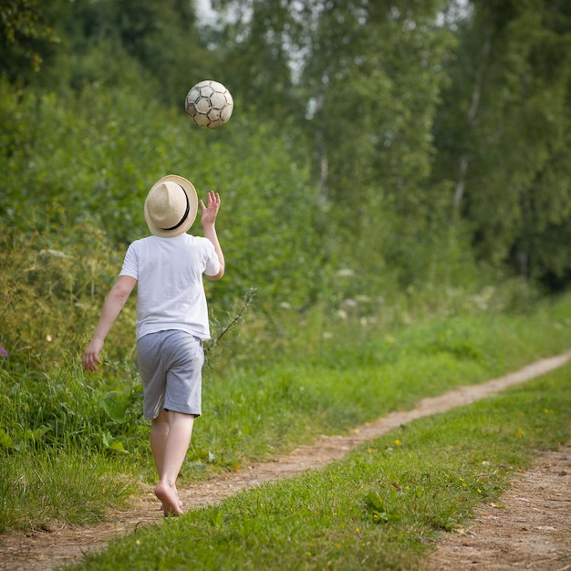 Widok z tyłu na chłopca w słomkowym kapeluszu spacerującego po wiejskiej drodze i bawiącego się piłką nożną
