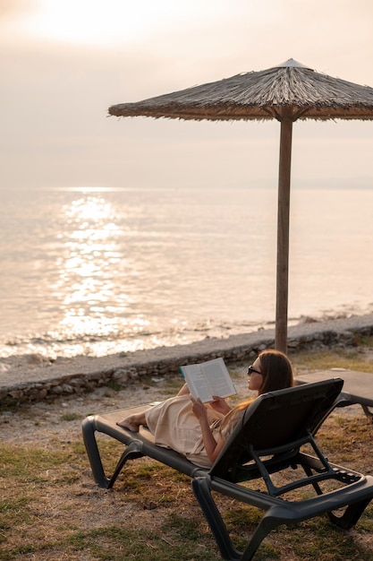 Widok z tyłu na damę w okularach przeciwsłonecznych spoczywającą na fotelu pod parasolem na plaży i czytającą powieść Turystka w letnim stroju ciesząca się wakacjami na wyspie Brac i spędzającą czas na wybrzeżu