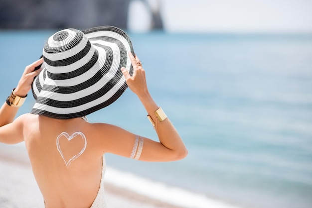 Zdjęcie widok z tyłu na piękną kobietę w pasiastym kapeluszu z kremem przeciwsłonecznym w kształcie serca na ramieniu, opalającą się na słynnej francuskiej plaży