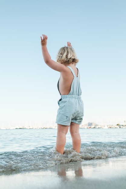 Widok z tyłu nierozpoznawalnego malucha w przypadkowych ubraniach, stojącego nad brzegiem morza i podnoszącego ręce, ciesząc się letnimi wakacjami
