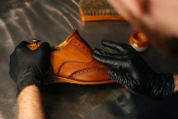 Zdjęcie widok z tyłu nierozpoznawalnego szewca w czarnych rękawiczkach pocierającego farbę o jasnobrązowe skórzane buty z...