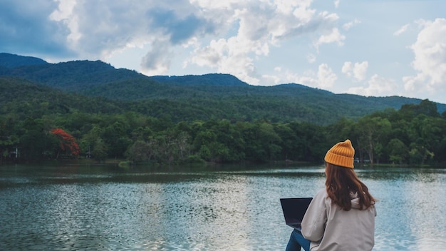 Widok z tyłu obrazu młodej kobiety korzystającej i pracującej na laptopie podczas podróży po górach i jeziorze