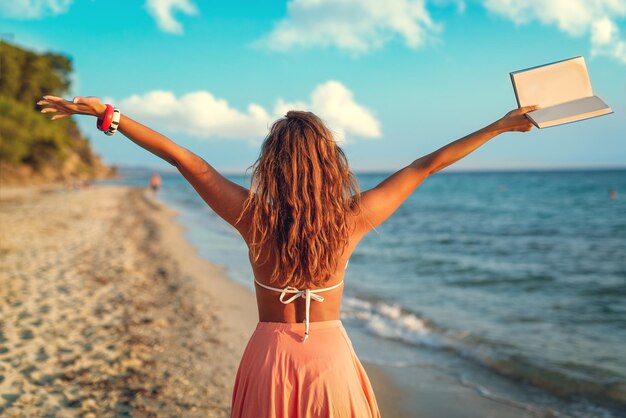 Widok z tyłu pięknej młodej kobiety, ciesząc się na plaży z książką w jej otwartych ramionach.