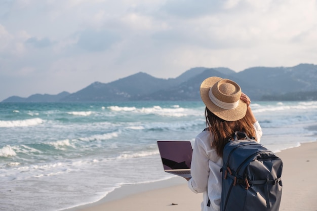 Widok z tyłu podróżniczki używającej i pracującej na laptopie podczas spaceru po plaży