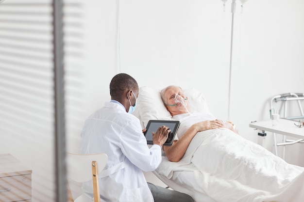 Widok z tyłu portret afroamerykańskiego lekarza za pomocą cyfrowego tabletu siedząc przez starszego pacjenta w szpitalnym łóżku, miejsce kopiowania