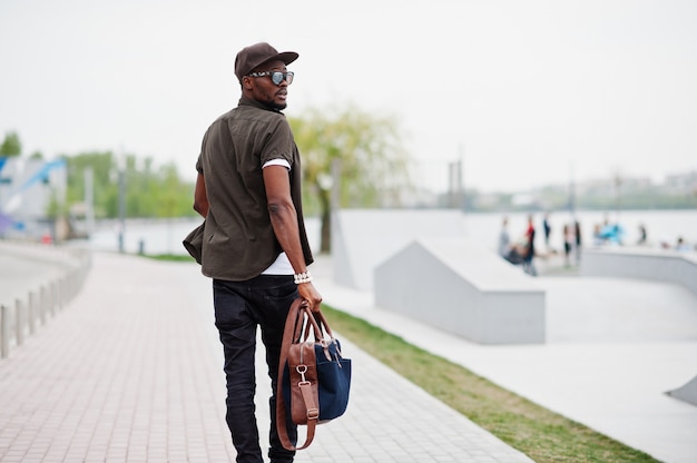 Widok z tyłu portret chodzenia stylowe African American Man nosić na okulary przeciwsłoneczne i czapkę z torebki na zewnątrz. Murzyn mody ulicznej.