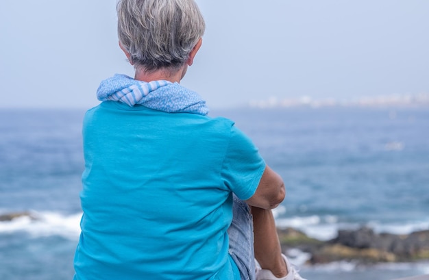 Widok z tyłu portret siwy starszy kobieta na sobie niebieską koszulkę i szalik, ciesząc się wakacje na świeżym powietrzu na morzu, odwracając się Zrelaksowany starsza pani siedzi na koncepcji emerytury klifu