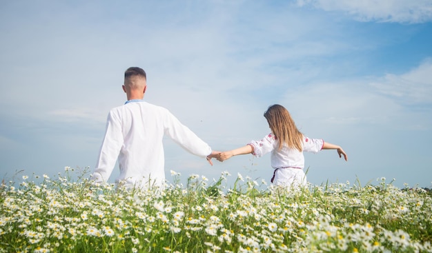 Widok z tyłu romantycznej pary w białych ubraniach, trzymającej się za ręce na polu z kwiatami rumianku, miłością i wolnością