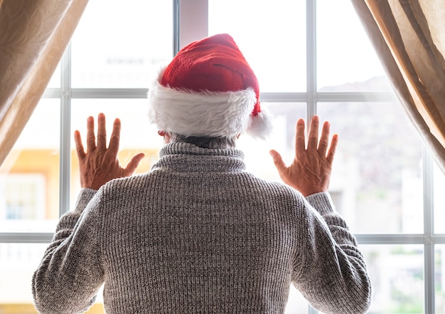 Widok z tyłu starszego mężczyzny w kapeluszu Świętego Mikołaja przebywającego w domu za oknem, patrząc na zewnątrz