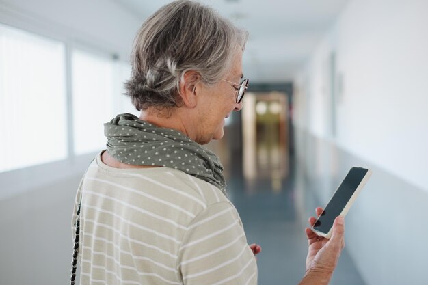 Widok z tyłu starszej białej kobiety chodzącej po korytarzu budynku podczas korzystania z telefonu Uśmiechnięta starsza pani czytająca wiadomość na smartfonie