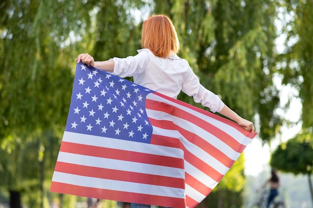 Widok z tyłu szczęśliwa młoda rudowłosa kobieta pozuje z flagą narodową USA stojącą na zewnątrz w letnim parku Pozytywna dziewczyna świętuje dzień niepodległości Stanów Zjednoczonych