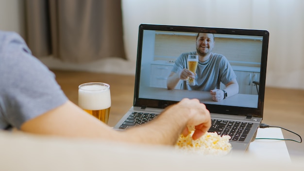 Widok z tyłu szczęśliwego człowieka na rozmowie wideo ze swoim przyjacielem pijącym piwo podczas izolacji koronawirusa.