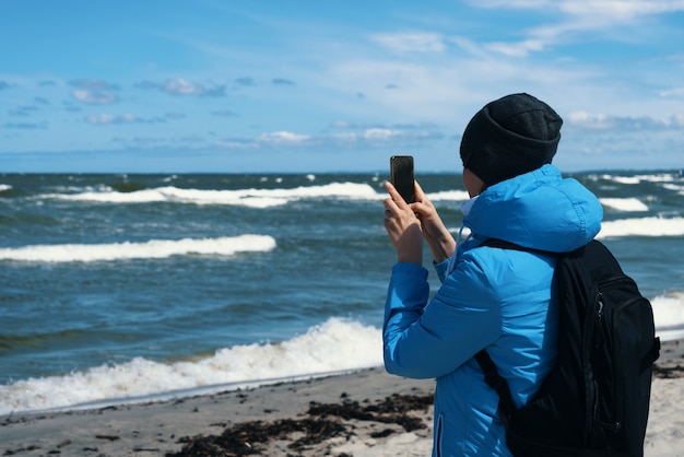 Widok z tyłu turysty dziewczyna robi zdjęcie aparatem cyfrowym telefonu komórkowego, stojąc nad morzem z falami