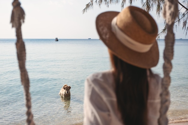 Widok z tyłu wizerunek kobiety siedzącej na huśtawce i patrzącej na psa bawiącego się wodą w morzu