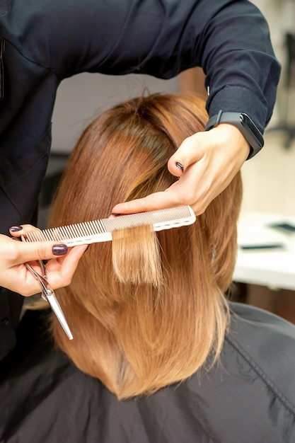Widok z tyłu z bliska fryzjer ścina czerwone lub brązowe włosy do młodej kobiety w salonie kosmetycznym. Strzyżenie w salonie fryzjerskim