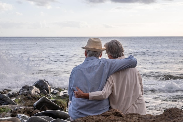 Widok z tyłu zrelaksowanej pary seniorów rasy kaukaskiej siedzącej na kamienistej plaży o zachodzie słońca podziwiający horyzont nad wodą Dwie starsze osoby o siwych włosach przytulające się z miłością