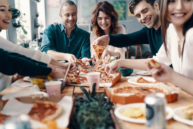 Więcej pizzy! Grupa młodych ludzi w strojach casualowych, którzy zbierają pizzę i uśmiechają się podczas kolacji w pomieszczeniu