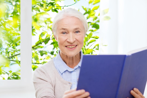 wiek, wolny czas i koncepcja ludzi - szczęśliwa uśmiechnięta starsza kobieta czytająca książkę w domu nad oknem i zielonym naturalnym tłem