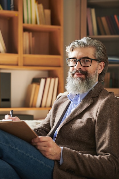 Wiekowy psycholog, odnoszący sukcesy w okularach i eleganckiej odzieży codziennej, patrzy na Ciebie na tle półek z profesjonalną literaturą