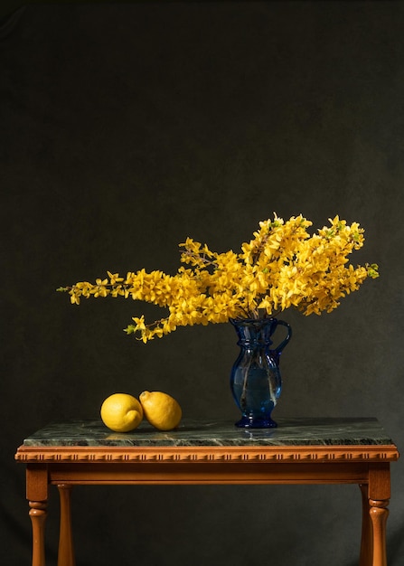 Wiele gałęzi z żółtymi kwiatami w niebieskim szklanym dzbanku na szarym tle Bukiet żółtych kwiatów na marmurowym stole jest wyłożony dwiema cytrynami Witamina C Dobre zdrowie