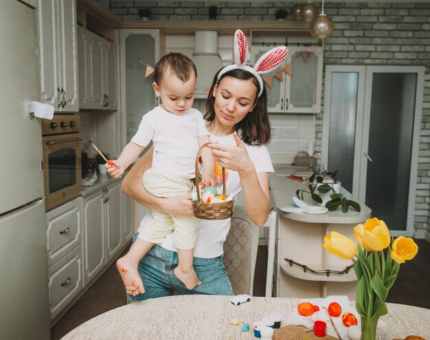 Wielkanocne Tradycje Rodzinne Kochająca Młoda Mama Uczy Dzieci Malowania Jajek Na Wielkanoc, Siedząc Razem Przy Kuchennym Stole