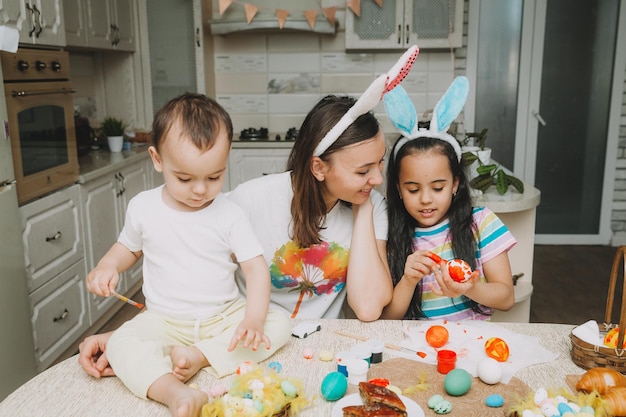 Wielkanocne Tradycje Rodzinne Kochająca Młoda Mama Uczy Dzieci Malowania Jajek Na Wielkanoc, Siedząc Razem Przy Kuchennym Stole