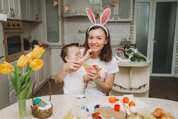 Wielkanocne tradycje rodzinne Kochająca młoda mama uczy dzieci malowania jajek na Wielkanoc, siedząc razem przy kuchennym stole