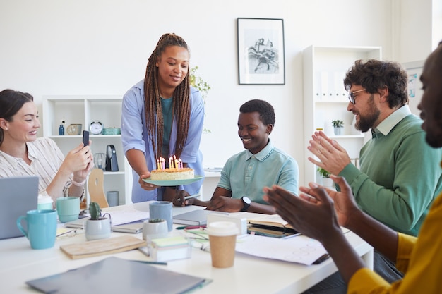 Wieloetniczna grupa ludzi obchodzi urodziny w biurze, skupia się na uśmiechniętej kobiecie przynoszącej tort młodemu Afroamerykaninowi