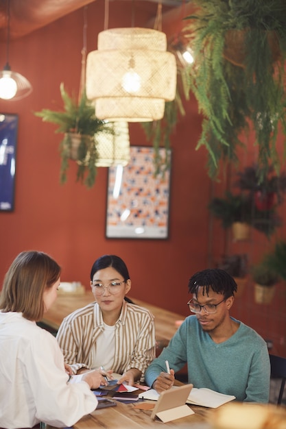 Wieloetniczna grupa młodych ludzi pracujących nad projektem, siedząc przy stole w kawiarni przed czerwoną ścianą