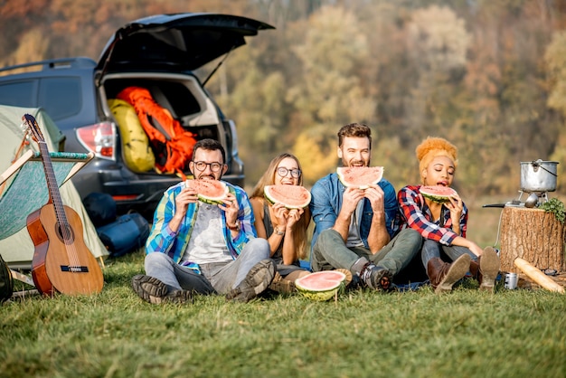 Wieloetniczna Grupa Przyjaciół Na Pikniku, Jedząca Arbuza, Siedząca W Rzędzie Na Kempingu Z Namiotem, Samochodem I Sprzętem Turystycznym W Pobliżu Jeziora