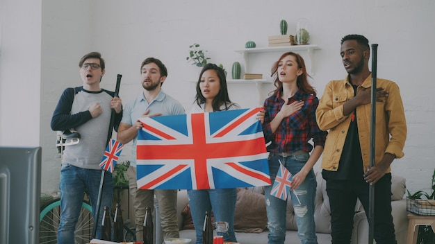Wieloetniczna grupa przyjaciół słuchająca i śpiewająca brytyjski hymn narodowy przed oglądaniem sportu