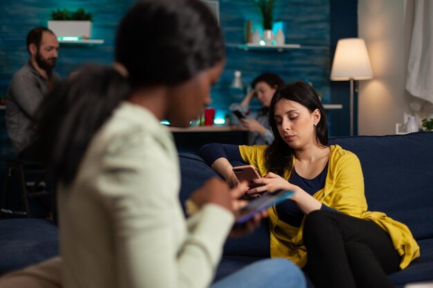 Wieloetniczni przyjaciele siedzący na kanapie, trzymający swojego nowoczesnego smartfona, surfują po Internecie, czytając wiadomości podczas relaksującej imprezy. Grupa ludzi cieszących się wspólnym spędzaniem czasu. koncepcja przyjaźni