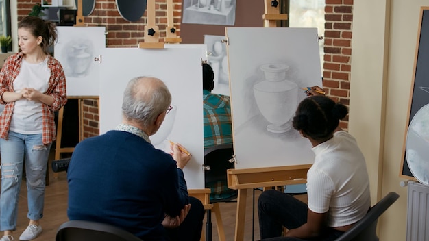 Wieloetniczny zespół ludzi uczący się techniki rysowania razem na zajęciach plastycznych. Starszy mężczyzna i młoda kobieta rozwijają umiejętności artystyczne i ćwiczą rysowanie modelu wazonu na płótnie.