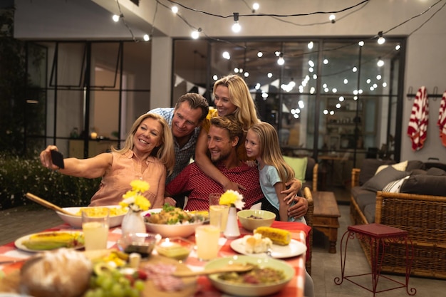 Wielopokoleniowa kaukaska rodzina spędza razem czas w domu, siedząc przy stole, nakładając jedzenie na talerze, jedząc razem, robiąc sobie selfie smartfonem