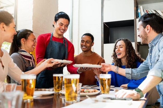 Wielorasowa grupa młodych przyjaciół jedzących i pijących piwo w restauracji Kelner serwujący pizzę