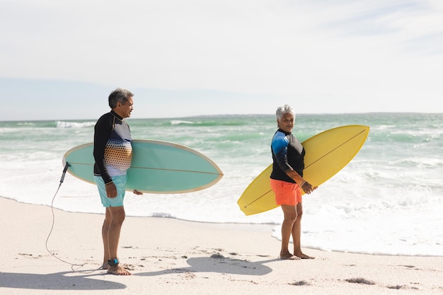 Wielorasowy starszy mężczyzna i kobieta niosący deski surfingowe na brzegu na plaży przed niebem w słoneczny dzień