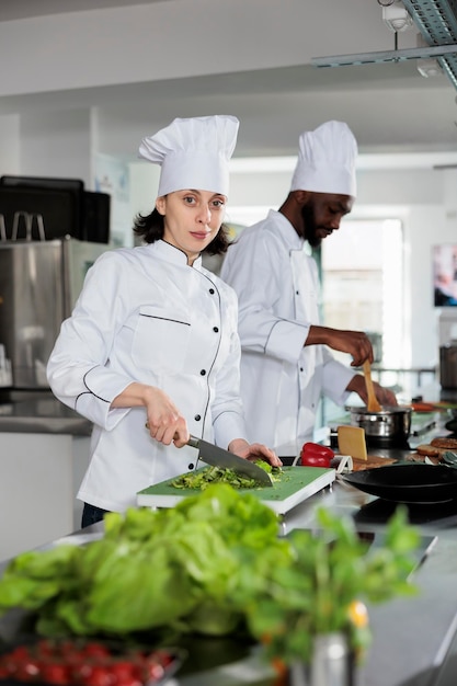 Wielorasowych pracowników przemysłu spożywczego stojących w profesjonalnej kuchni restauracji podczas gotowania smacznego dania. Przekonany szef kuchni krojenia świeżych warzyw do dekoracji, uśmiechając się do kamery.