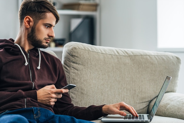 Wielozadaniowość. Widok z boku skoncentrowanego młodego mężczyzny korzystającego z laptopa i trzymającego smartfona siedzącego na kanapie w domu