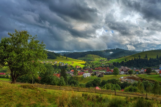 Wieś w górach, na tle zielonych zboczy i lasów, niebo z chmurami burzowymi