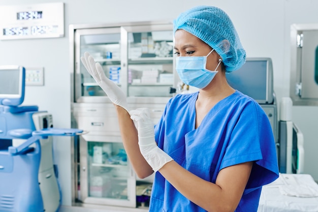 Wietnamska chirurg kobieta w masce medycznej, zakładająca gumowe rękawiczki i przygotowująca się do operacji
