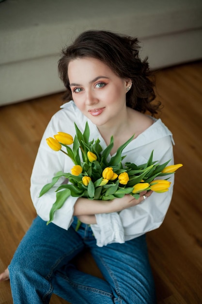 Wiosenny portret młodej pięknej kobiety o niebieskich oczach i kręconych włosach z bukietem żółtych tulipanów we wnętrzu domu Szczęśliwy model w dżinsach i białej koszuli Dzień Matki