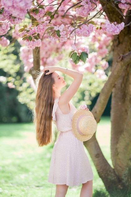 Wiosenny portret młodej pięknej kobiety w różowych kwiatach Młoda piękna modelka z długimi włosami w wiklinowym kapeluszu i lekkiej sukience w pobliżu kwiatów wiśni