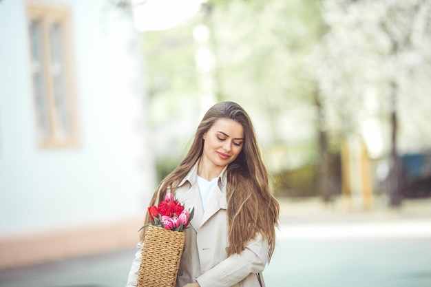 Wiosenny portret młodej pięknej szczęśliwej kobiety w wieku 28 lat z długimi zadbanymi włosami trzyma w rękach wiklinową torbę z bukietem tulipanów na ulicy miasta Stylowy model w trenczu