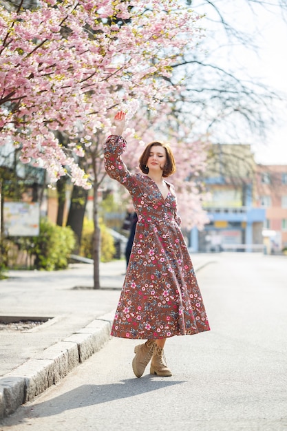 Wiosną kobieta spaceruje po kwitnącej ulicy z drzewami sakura. Dziewczyna w długiej jedwabnej eleganckiej sukience w stylu vintage spaceruje wśród kwitnących drzew