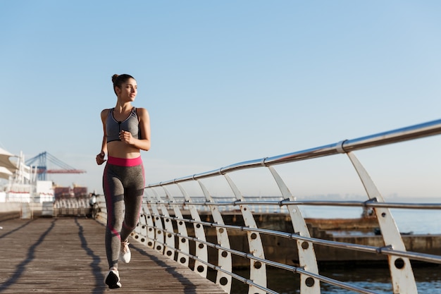 Wizerunek atrakcyjnej kobiety lekkoatletycznego w sportowej joggingu rano, patrząc na morze i uśmiechnięty