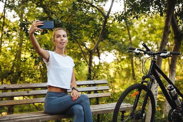 Wizerunek Europejskiej kobiety siedzącej na ławce w parku z rowerem i robienia zdjęć selfie na telefon komórkowy