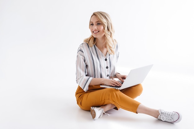 Wizerunek ładnej uśmiechniętej kobiety noszącej ubranie, wpisując na laptopie i patrząc na bok, siedząc na podłodze samodzielnie nad białą ścianą