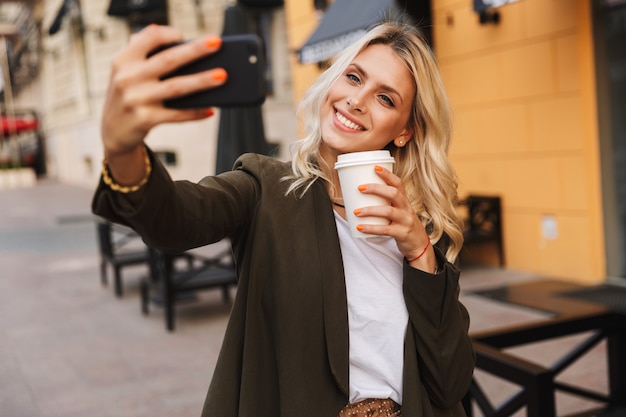 Wizerunek Pięknej Kobiety Trzymającej Kawę Na Wynos W Papierowym Kubku I Biorąc Selfie, Spacerując Ulicą Miasta