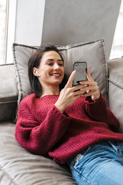 Wizerunek pięknej kobiety w pomieszczeniu w domu na kanapie przy użyciu telefonu komórkowego.